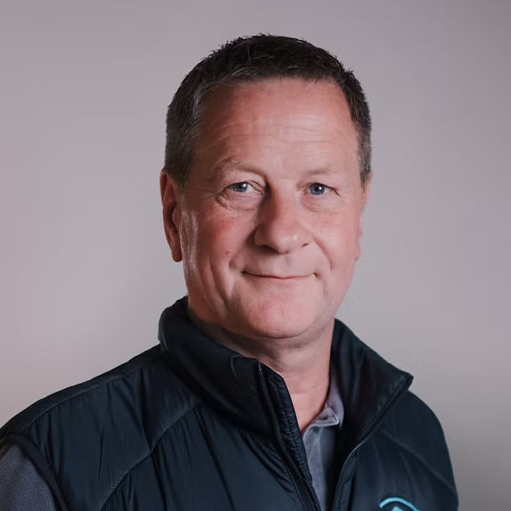 A professional headshot of a man with short brown hair, wearing a black zip-up vest over a grey collared shirt against a light grey background.