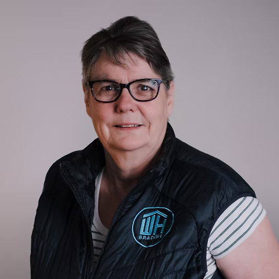 A professional headshot of a woman with short dark hair and glasses, wearing a striped shirt under a black vest featuring a teal "WH BRADING" logo.