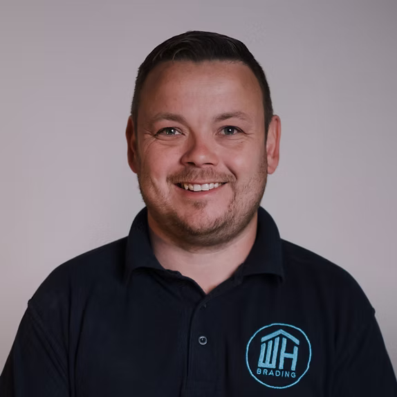 A professional headshot of a smiling man with short brown hair and a light beard, wearing a dark blue polo shirt featuring a light blue "WH BRADING" logo.