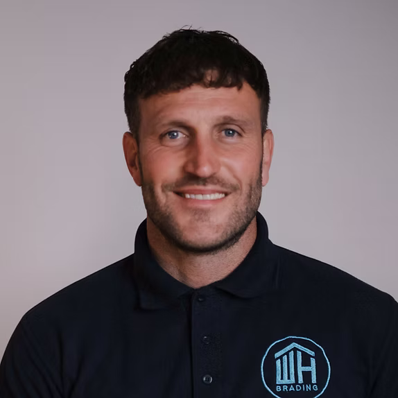 A professional headshot of a smiling man with short dark hair, wearing a dark blue polo shirt featuring a light blue "WH BRADING" logo.