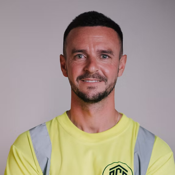 A professional headshot of a smiling man with short dark hair and a beard, wearing a bright yellow high-visibility work shirt with grey reflective stripes.