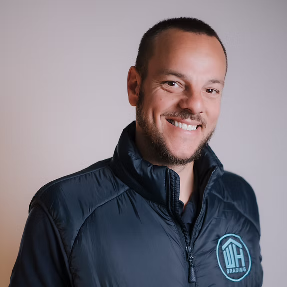 A professional headshot of a brightly smiling man with short brown hair and a beard, wearing a black zip-up vest featuring a light blue "WH BRADING" logo.