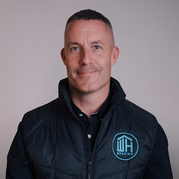 A professional headshot of a smiling man with short dark hair, wearing a black zip-up vest with a light blue "WH BRADING" logo over a dark shirt.